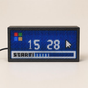May include: A rectangular black digital clock with a dot matrix display. The screen shows the time 15:28 in white, a colorful four-square icon, a white cursor arrow, and a loading bar with the text START in white pixels on a blue background.