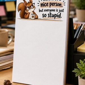 May include: A white notepad featuring a cartoon illustration of a brown squirrel wearing black glasses and holding a small mug. The text reads, I want to be a nice person.. but everyone is just so stupid. The notepad has a large blank area below.