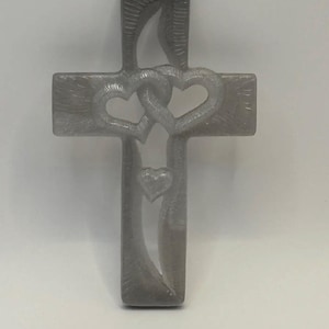 May include: A grey cross with a smooth, semi-translucent finish featuring two interlocking hearts at the centre and a smaller heart positioned below them. The surface of the cross displays subtle, textured, linear patterns throughout its structure.