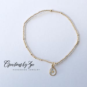May include: A delicate gold bracelet with tiny beads and a teardrop-shaped charm containing a small gold speck. The bracelet is on a white background with the text "Creations by Zee Handmade Jewelry".