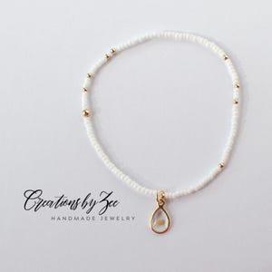 May include: A delicate white beaded bracelet with a gold teardrop charm. The charm has a small, clear stone inside.