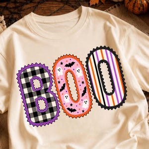 May include: A cream-colored t-shirt featuring the word BOO in large letters. The B is black and white buffalo plaid, the O is pink with black bats and white ghosts, and the final O has vertical stripes in orange, purple, black, and white.
