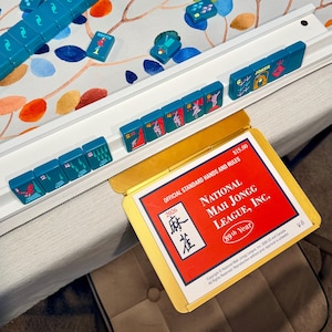 May include: A white plastic Mah Jongg tile rack holding teal tiles with colorful graphics, including numbers and symbols. A yellow card holder displays the 2026 National Mah Jongg League, Inc. Official Standard Hands and Rules card with red background.