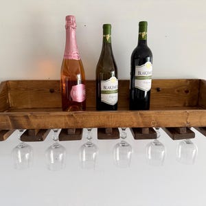May include: A rectangular wall-mounted wooden wine rack with a dark brown finish. The top shelf holds three glass bottles, while the bottom section features six slots designed to hang wine glasses upside down by their stems.