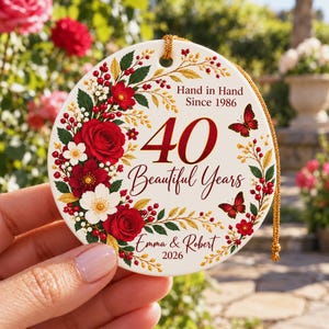 May include: A round white ceramic ornament featuring a floral wreath of red roses, white flowers, and red berries. The text reads Hand in Hand Since 1986, 40 Beautiful Years, Emma & Robert, and 2026. Two red butterflies are printed on the surface.