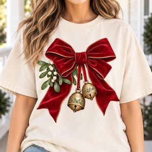 Puede incluir: Una camiseta de manga corta color crema con un gráfico central grande de un lazo de terciopelo rojo intenso con bordes dorados. El lazo incluye ramas de muérdago verde con bayas blancas y dos cascabeles dorados colgantes con grabados de acebo.