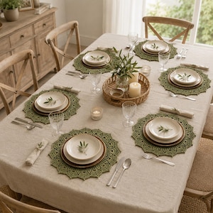 May include: A dining table set for six with a beige linen tablecloth, featuring round olive green crocheted placemats, white ceramic plates, clear glassware, stainless steel cutlery, linen napkins, and a central woven basket with candles.