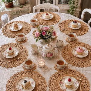 May include: A round dining table set for six with beige crocheted placemats, floral porcelain teacups, saucers, and plates. The table features a central floral teapot, a creamer, a sugar bowl, a vase of pink roses, and individual cupcakes or cake slices.