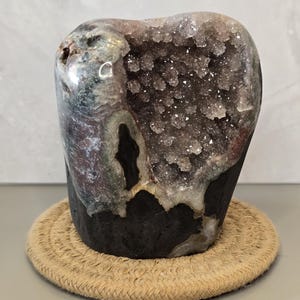 May include: A natural geode specimen featuring a dark, smooth exterior base that transitions into a hollow center filled with small, sparkling purple amethyst crystals. The stone has a rounded, irregular shape with varied tones of brown and gray.