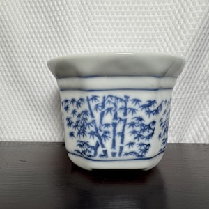 May include: A white ceramic planter with an octagonal shape and a blue hand-painted bamboo forest pattern. The vessel features a flared rim and a smooth, glossy finish, showcasing detailed blue botanical illustrations encircling the exterior.