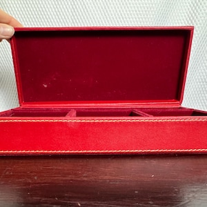 May include: A rectangular red storage box with a hinged lid, featuring a smooth exterior finish and contrasting light-colored stitching along the edges. The interior is lined with a deep red velvet fabric and divided into three separate compartments.