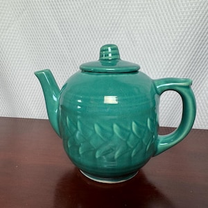 May include: A teal ceramic teapot with a glossy finish and a removable lid. The body of the teapot features a raised, repeating leaf-like pattern encircling the center. It has a curved spout on the left and a sturdy handle on the right side.