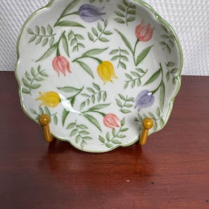 May include: A white ceramic bowl with a scalloped edge and a green rim, featuring a painted pattern of yellow, pink, and purple tulips interspersed with green leafy stems. The bowl is displayed on a small, two-pronged amber-colored stand.
