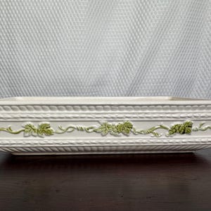 May include: A rectangular off-white ceramic baking dish featuring a raised relief pattern of green grape vines and leaves along the sides, complemented by a textured border of horizontal lines and small repeating indentations.