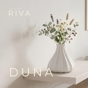 Può includere: Un vaso in ceramica bianca con base bombata e scanalata contenente un bouquet di lavanda essiccata, margherite bianche, foglie di eucalipto e piume d'erba. Il testo COLLEZIONE RIVA DUNA è visibile negli angoli dell'immagine.