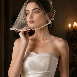 May include: A sheer, short bridal veil featuring a delicate trim of white pearls and thin, dangling silver-toned chains. The veil is crafted from fine, translucent tulle fabric, creating a soft, layered effect that frames the head and shoulders.