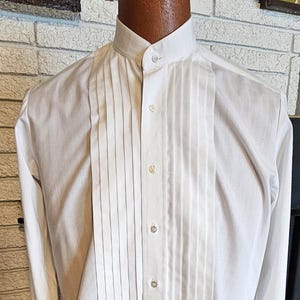 Puede incluir: Una camisa blanca formal con un frente de peto plisado, mangas largas y cuello clásico. La camisa tiene cierre de botones y se muestra en un maniquí. Adecuada para eventos formales.