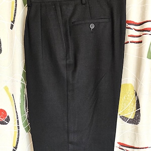 Puede incluir: Pantalones negros con cierre de botón y parte delantera plisada. Los pantalones cuelgan de una percha sobre un fondo estampado. El dobladillo de los pantalones está vuelto.