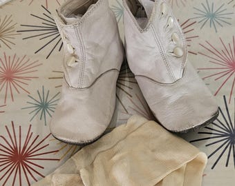 May include: A pair of off-white vintage baby boots with button closures and a pair of matching stockings. The items are placed on a patterned surface with starburst designs in various colors.