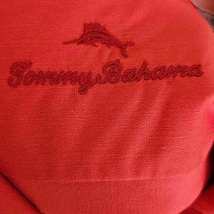 May include: Red collared shirt with a white stitching detail around the collar. The shirt has an embroidered logo of a marlin fish and the text "Tommy Bahama" in red thread.