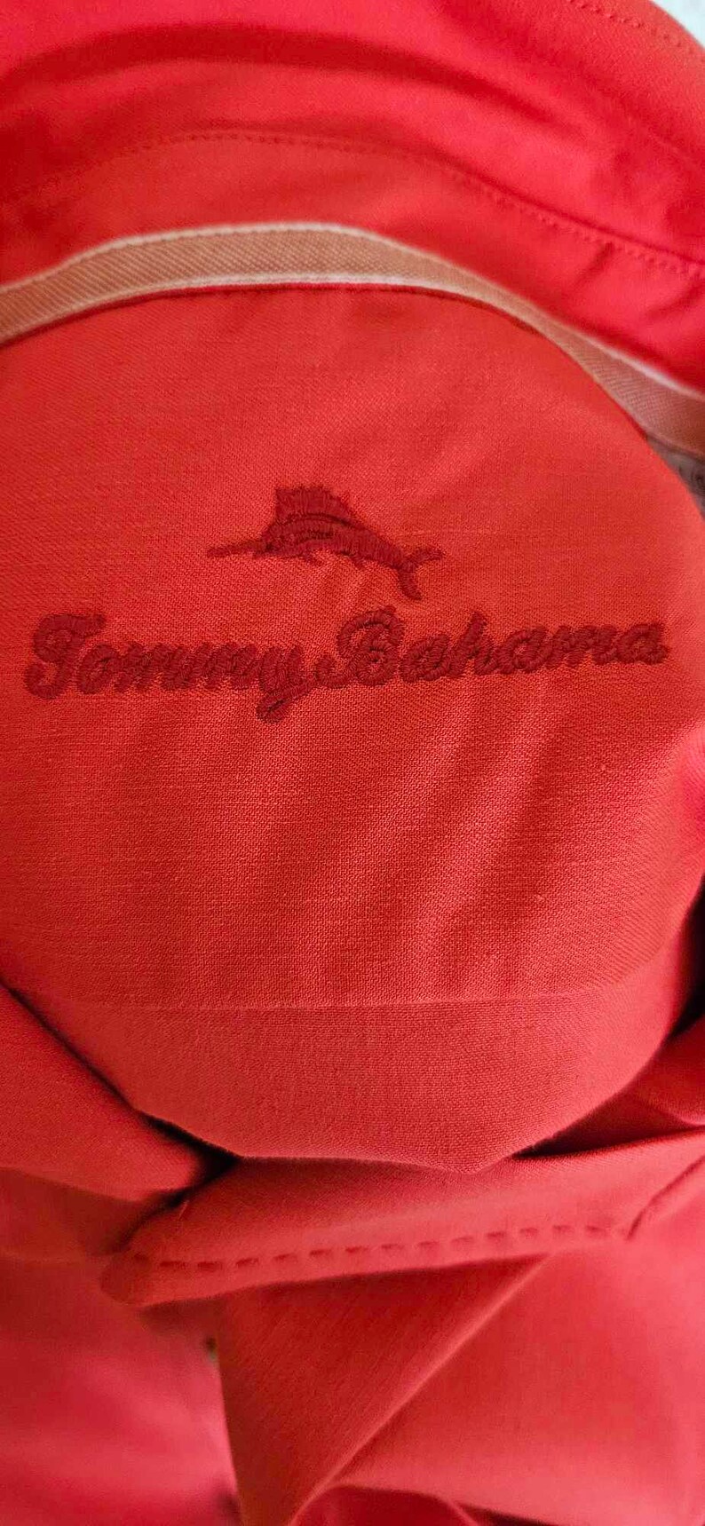 May include: Red collared shirt with a white stitching detail around the collar. The shirt has an embroidered logo of a marlin fish and the text "Tommy Bahama" in red thread.