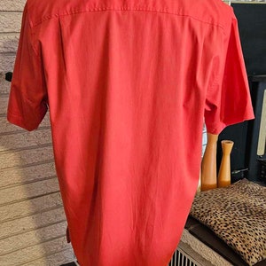 May include: A red short-sleeved button-down shirt on a mannequin. The shirt is made of a smooth, lightweight fabric.