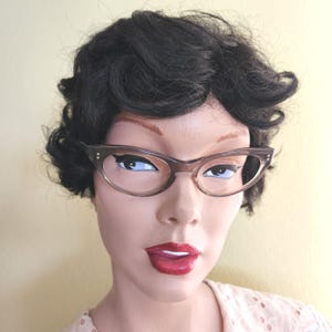 May include: A mannequin head with dark curly hair and brown cat-eye glasses. The glasses have a light brown frame with small silver accents on the temples. The mannequin has red lipstick and a light pink top.