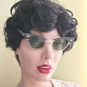 May include: Clear cat-eye sunglasses with black and white polka dot detailing on the frames. The sunglasses have a retro style with a clear frame and green-tinted lenses. The sunglasses are a vintage accessory.