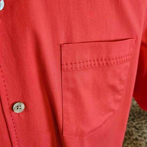 May include: A red short-sleeved button-down shirt with a single chest pocket and four buttons. The shirt has a subtle texture and a classic design.