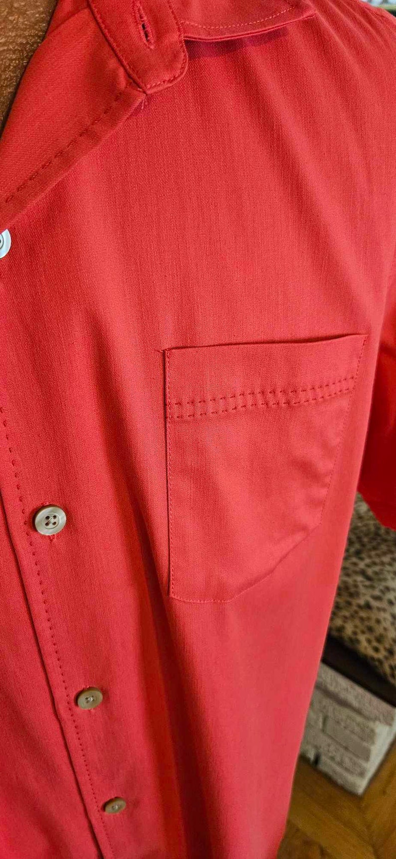 May include: A red short-sleeved button-down shirt with a single chest pocket and four buttons. The shirt has a subtle texture and a classic design.