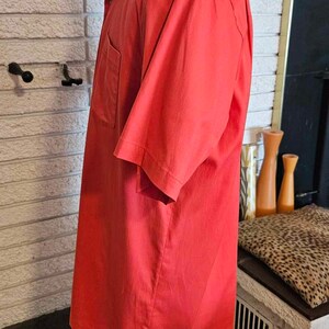 May include: A red short-sleeved shirt with a button-down collar and a single pocket on the left side. The shirt is on a mannequin and is displayed against a white brick wall.