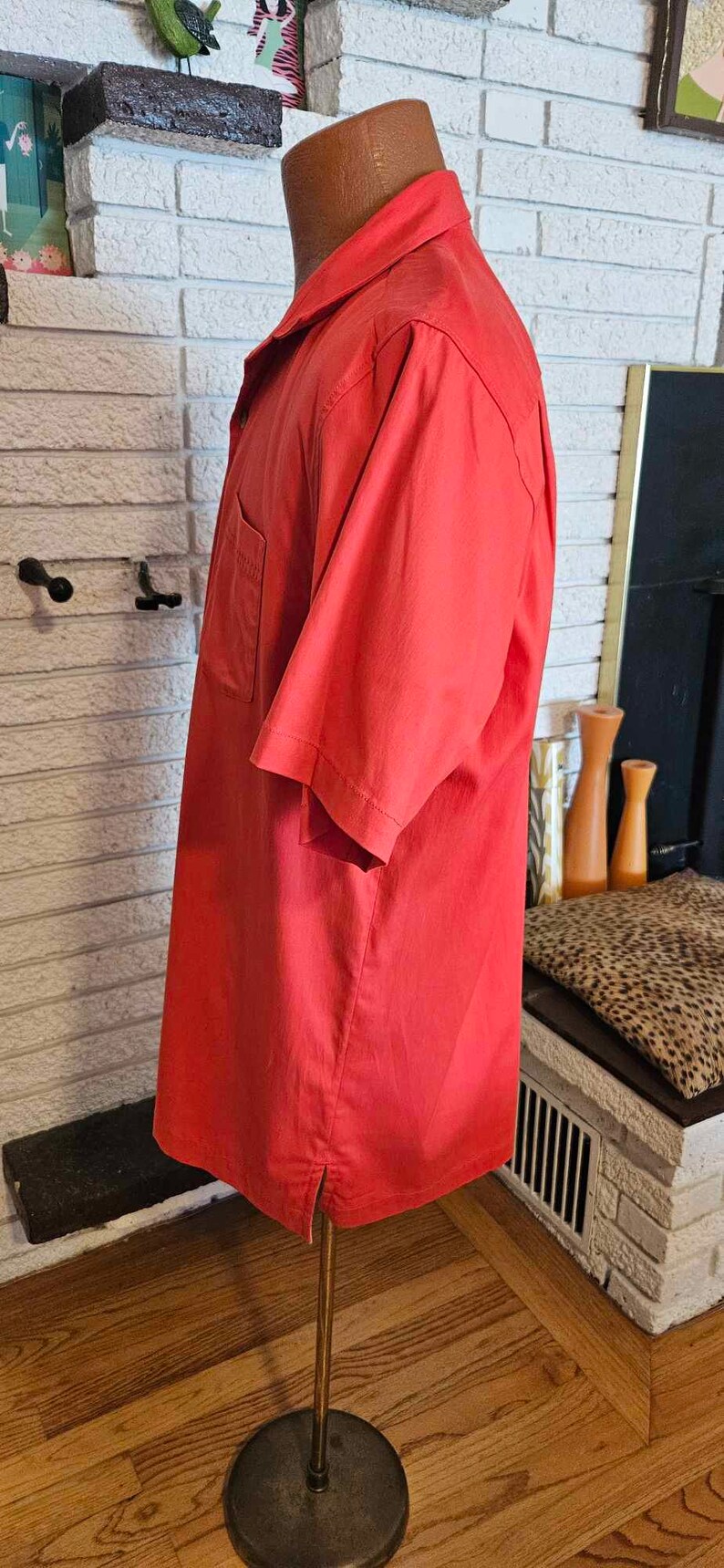 May include: A red short-sleeved shirt with a button-down collar and a single pocket on the left side. The shirt is on a mannequin and is displayed against a white brick wall.