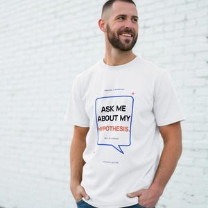 May include: A white short-sleeved t-shirt featuring a blue speech bubble graphic containing the black text "ASK ME ABOUT MY HYPOTHESIS." with the word "HYPOTHESIS." in orange. Additional small text includes "TEEXCON / RETRO DOG" and "CURIOSITY WELCOME."