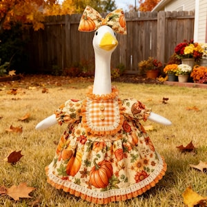 May include: A white plush goose figurine wearing a dress with a pumpkin and autumn leaf print, featuring a matching bow on its head, an orange and white gingham bib with two wooden buttons, and orange lace trim along the hem and collar.