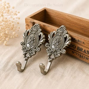May include: Two matching wall hooks made of antique silver-toned metal. Each hook features an oval center medallion with a raised floral relief pattern, surrounded by ornate scrollwork and a curved hook at the bottom with a circular mounting hole.