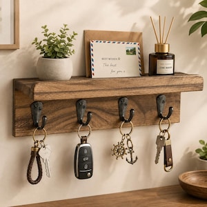 May include: A wall-mounted wooden shelf with four dark metal hooks. The shelf holds a small potted green plant, a greeting card with the text "BEST WISHES The best for you", and a glass bottle with reed diffusers. Four keychains hang from the hooks.