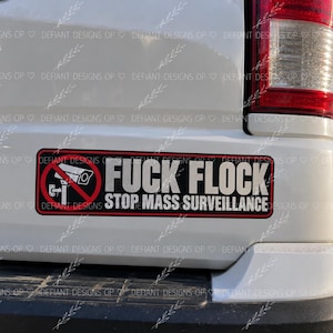 May include: A rectangular bumper sticker with a black background and a red border. On the left is a red circle with a diagonal line over a white security camera icon. The text reads FUCK FLOCK STOP MASS SURVEILLANCE in bold white letters.