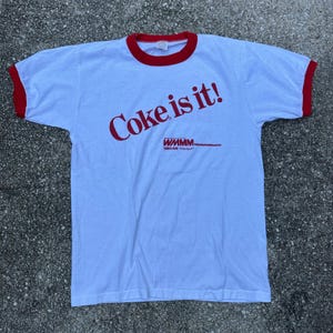 May include: A white ringer t-shirt with red trim on the collar and sleeves. The front features the red text Coke is it! printed diagonally, with the text WMMM 1260AM Westport printed in red below the main slogan.