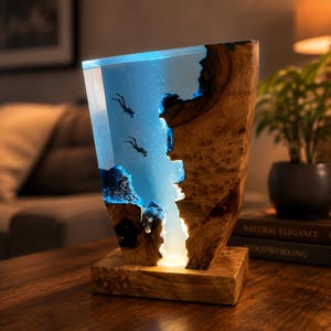 Könnte beinhalten: Eine dekorative Lampe aus einem vertikalen Stück Naturholz mit Baumkante, kombiniert mit durchscheinendem blauem Harz. Der Harzbereich enthält zwei kleine Taucherfiguren und wird von unten beleuchtet, was einen leuchtenden Unterwassereffekt erzeugt.