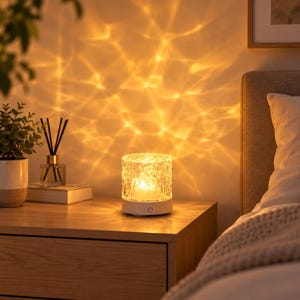 May include: A cylindrical table lamp with a textured, clear glass shade that emits a warm, golden light, casting intricate, rippling patterns onto the wall. The lamp features a white base with a power button icon on the front surface.