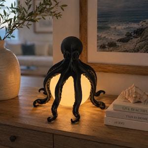 Könnte beinhalten: Eine schwarze, oktopusförmige Lampe mit einer zentralen, leuchtenden, kugelförmigen Lichtquelle. Der Oktopus hat acht geschwungene Tentakel, die nach außen ragen und die beleuchtete Kugel umgeben. Die Lampe steht auf einer Holzoberfläche.