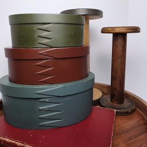 May include: A stack of three oval wooden pantry boxes in olive green, deep red, and muted blue, each featuring finger-jointed sides with small metal tacks. Beside the boxes stands a tall, dark brown wooden spool with a flat circular top.