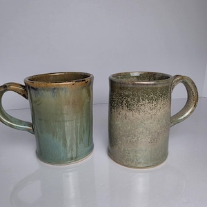 May include: Two ceramic mugs with handles. The left mug features a smooth, glossy glaze in shades of sage green and brown with vertical drip patterns. The right mug has a textured, speckled finish in muted olive green and beige tones.