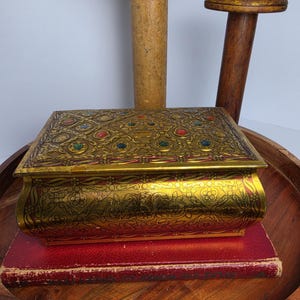 May include: A rectangular gold-toned metal box featuring an intricate embossed floral pattern and inlaid colorful faux gemstones on the lid. The box sits atop a red hardcover book, which is placed on a dark wooden tray with two wooden spools.