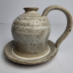 May include: A ceramic butter bell with a speckled beige and off-white glaze. The piece features a rounded body with a small opening at the top, a wide circular base, and a single curved handle attached to the side of the main vessel body.