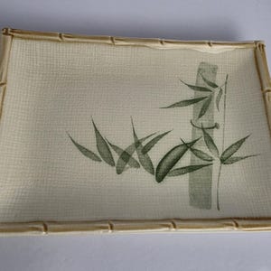 May include: A rectangular ceramic serving tray with a cream-colored surface featuring a subtle grid texture. The rim is molded to resemble bamboo stalks in a light tan glaze. A green painted illustration of bamboo stalks and leaves is centered.