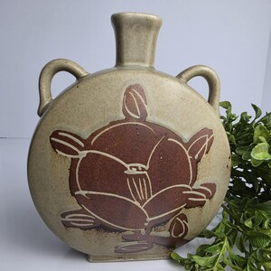 Puede incluir: Una petaca de cerámica redonda de color beige con dos asas laterales y un cuello estrecho. La parte frontal presenta un gran diseño floral de color marrón oscuro incrustado con líneas talladas. El recipiente tiene una base plana y un acabado mate suave, mostrando un contraste entre el cuerpo beige claro y el motivo central marrón oscuro.