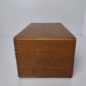 May include: A rectangular wooden box with a visible brown wood grain pattern. The box features interlocking finger joints at the corners, connecting the front panel to the side panels. The surface shows a natural texture with horizontal wood grain.