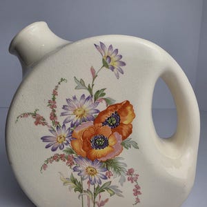 May include: A cream-colored ceramic pitcher with a rounded, asymmetrical shape and a side handle. The surface features a fine crackle glaze finish and a central floral decal depicting orange poppies and purple daisies with green foliage.