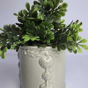 Könnte beinhalten: Ein hellgrauer Keramikübertopf in zylindrischer Form mit geprägten Herz- und Topiary-Motiven auf der Außenfläche sowie einem dekorativen Rand an der Ober- und Unterseite, bepflanzt mit einer üppigen, künstlichen grünen Blattpflanze.